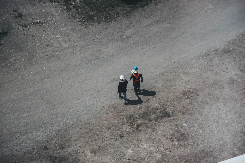 Two construction workers walking on a gravel road with safety gear and helmets, seen from above.