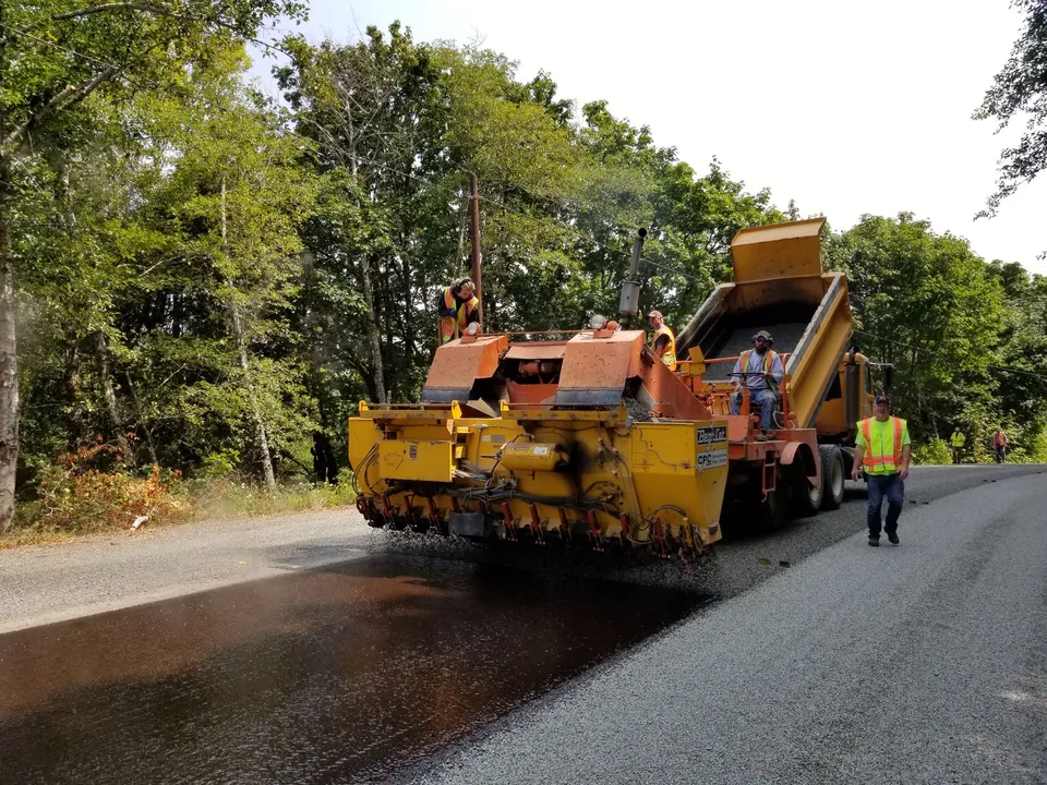 Road paving in Oregon forest with workers and machinery. Summer construction scene outdoors.